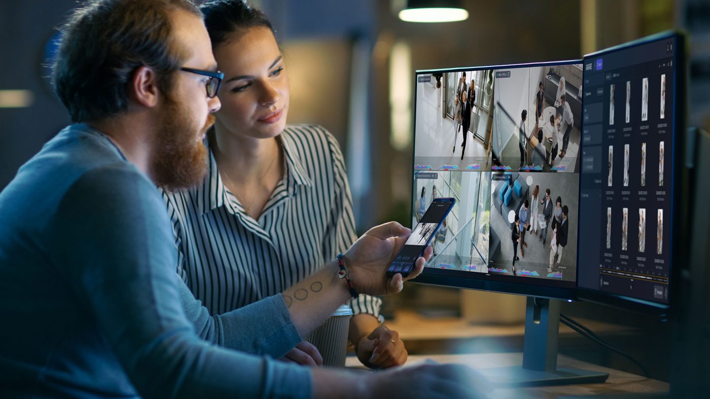 A man and a woman are looking at a multi-monitor display showing multiple video feeds from what appears to be a security or surveillance system. The man is holding a smartphone, which also shows a video feed. The monitors display live footage and what looks like a list of event logs or video clips on the far right screen. The man has a beard and glasses, and the woman is looking intently at the screens. The setting appears to be an office or control room at night.