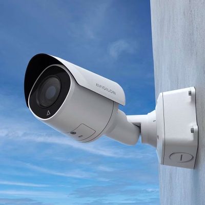 A white Avigilon H6SL bullet security camera mounted on a wall against a blue sky.
