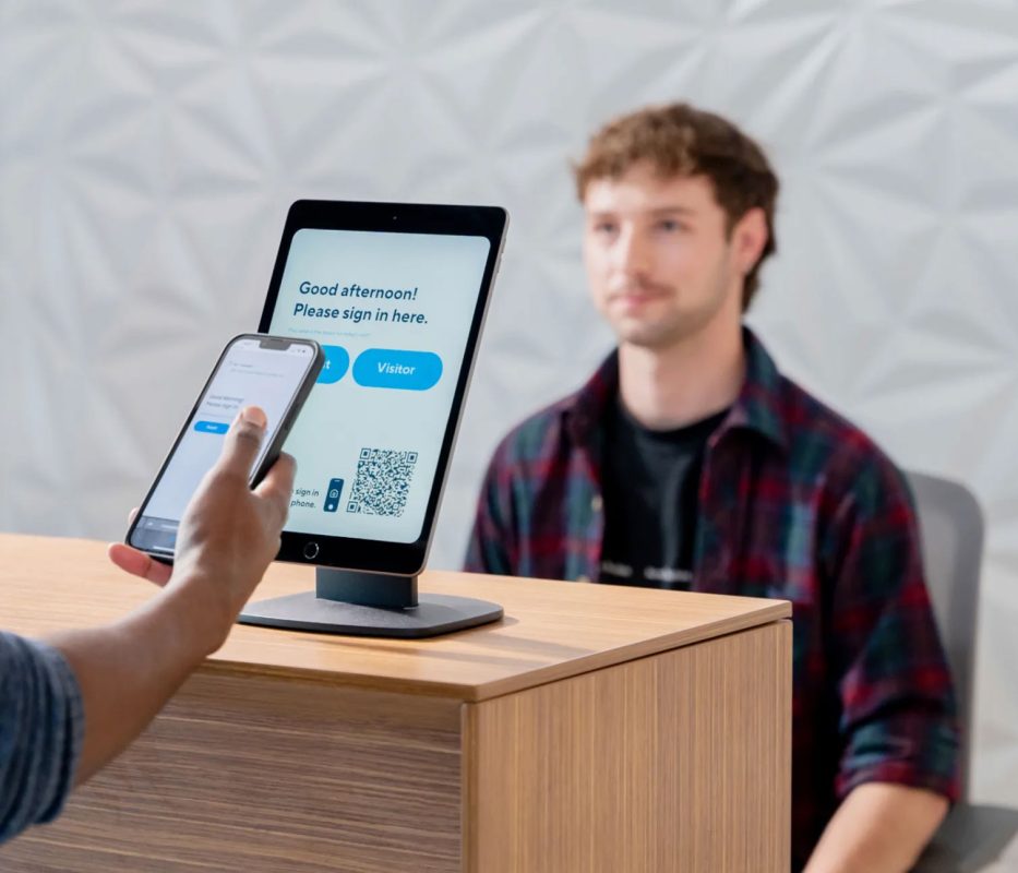 A person using a smartphone to check in on a tablet device with a QR code and a "Visitor" button on the screen. A man is seated in the background.