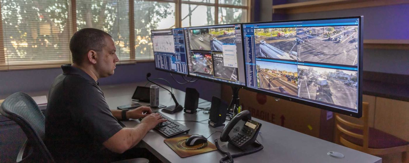 A man sits at a desk with multiple monitors displaying surveillance feeds and data, likely in a police or security control room.