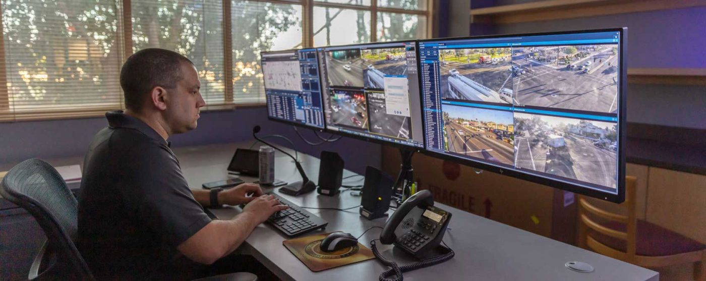 A man, who is an operator at the Glendale Police Department's Real Time Crime Center, sits at a desk with multiple monitors displaying live camera feeds of traffic intersections and other data.