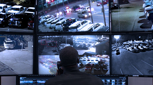 a security guard or operator monitoring multiple security camera feeds on a bank of screens. The feeds display various views, including a parking lot at night, a street with traffic, and what appears to be a car dealership or a similar commercial area. The person is sitting with their back to the camera, focused on the monitors.